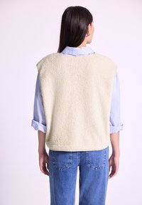 Gilet en polaire beige avec une finition texturée, design sans manches, bords arrondis, porté sur une chemise à manches longues bleu clair, associé à un jean en denim.