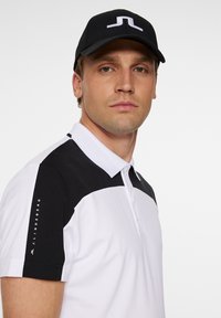 Svart och vit polo shirt med svart krage och axelpartier. Kappen har en vit logotyp på svart bakgrund. Enkel design.