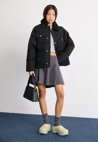 Padded crna jakna s krznim ovratnikom, sivi crop top, siva naberena suknja i svijetlozelene klompe. Crna torba s oblikovanim dizajnom.