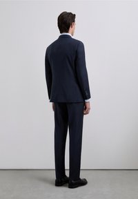Traje a medida azul marino con blazer estructurado con abertura trasera única, pantalones de corte recto y zapatos de cuero negro, visto desde atrás.