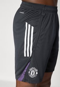 Sorte sportsshorts med hvid tre-stripes design på siden, lilla akcentpanel og Manchester United-logo på kanten.
