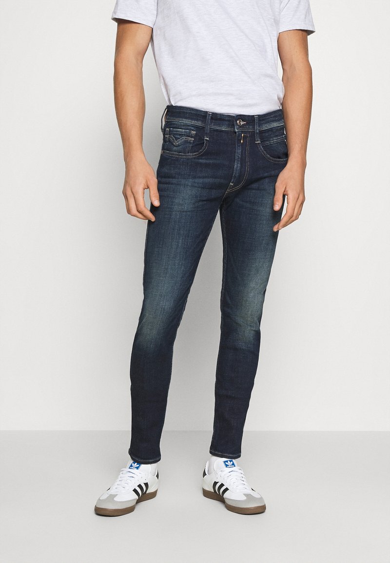 Jean slim bleu foncé en denim, avec une taille mi-haute, un design à cinq poches et de légères marques d'usure. Associé à des baskets blanches.