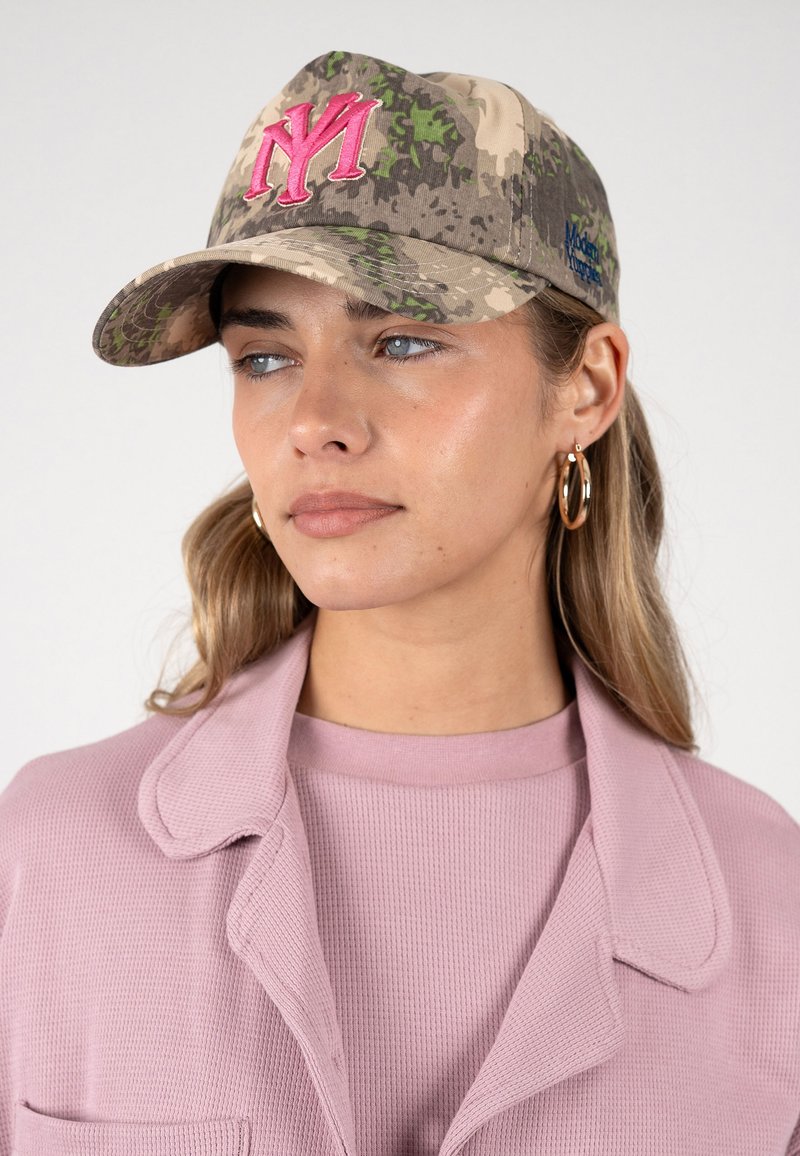 Femme portant une casquette camouflage avec un lettrage "NY" brodé rose, des boucles d'oreilles créoles dorées et une veste rose texturée par-dessus un haut assorti.