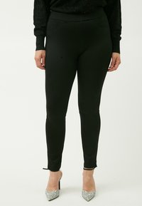 Zwarte legging met een zachte textuur, hoge tailleband en zijzakken. Verfraaid met kleine strikjes bij de enkel en gecombineerd met glinsterende hakken.