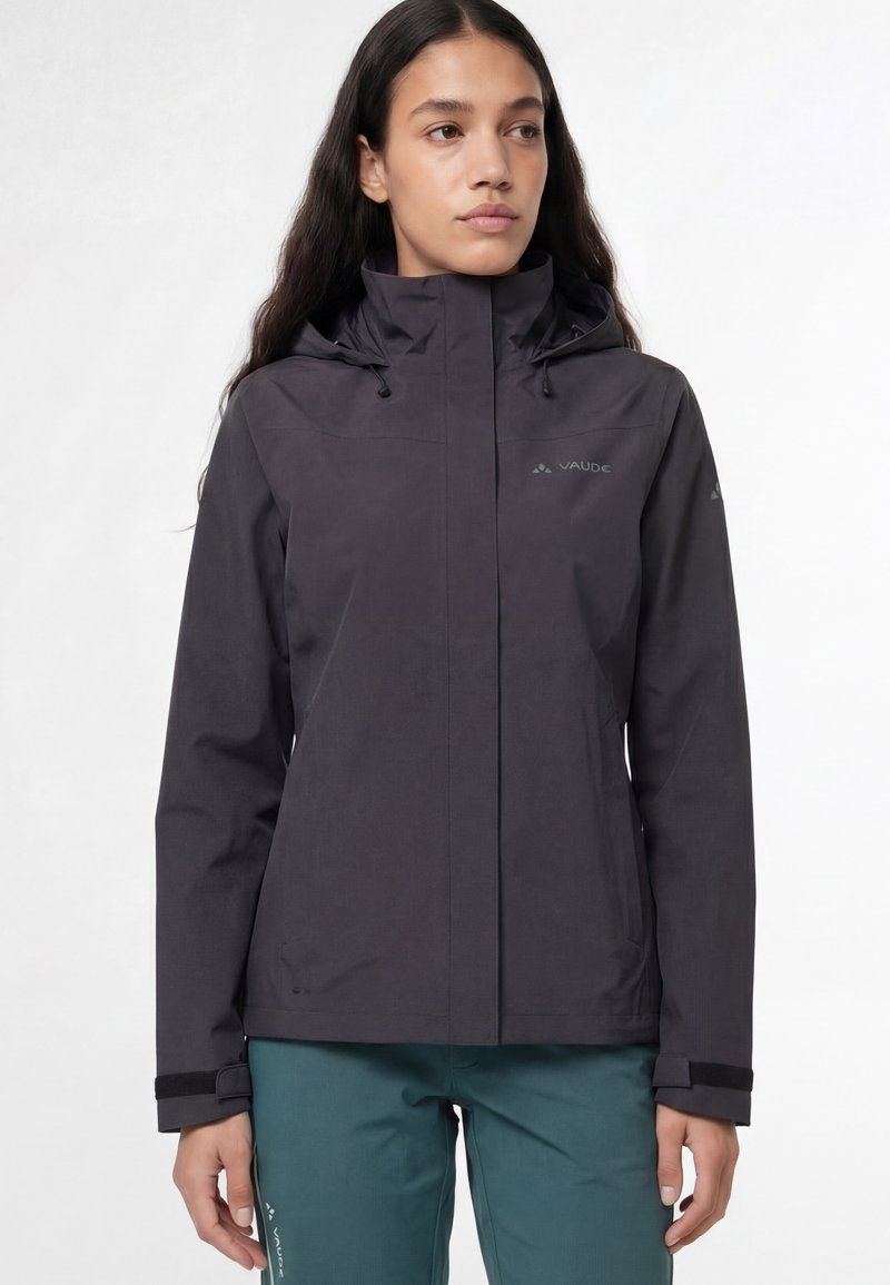 Donna che indossa una giacca impermeabile nera con cappuccio e pantaloni da esterno verde acqua, in piedi contro uno sfondo chiaro uniforme.