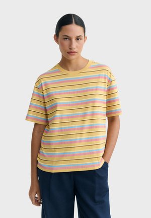 Młoda kobieta z ciemnymi włosami w paskowaną żółtą, różową i niebieską koszulkę z krótkim rękawem oraz granatowe spodnie, stoi z jedną ręką w kieszeni.