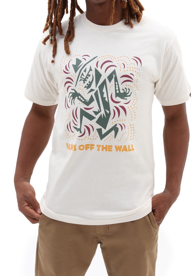 Personne portant un t-shirt blanc Vans avec une figure abstraite dentelée aux bras étendus et le texte « Vans Off The Wall » en orange.