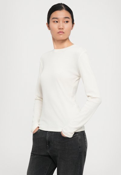 Kvinna med mörkt hår i en vit långärmad topp och svarta jeans står med händerna i fickorna mot en enfärgad bakgrund.