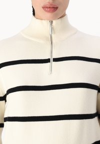 Cremfarbener Pullover mit schwarzen horizontalen Streifen, ausgestattet mit einem hohen Kragen und einem halben Reißverschluss. Hergestellt aus weichem, strukturiertem Material.