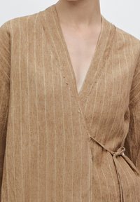 Haut en lin marron avec de fines rayures verticales, décolleté en V et taille nouée, présentant une finition texturée et des manches amples.