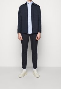 Giacca di cotone blu navy con colletto e chiusura a bottoni, camicia azzurra chiara con bottoni, pantaloni scuri e sneakers color crema.