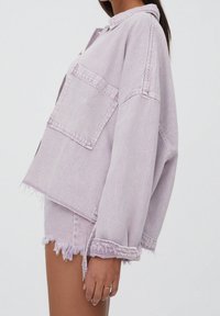Mujer con una chaqueta de mezclilla ligera de gran tamaño en color púrpura claro, con un gran bolsillo delantero, y shorts de mezclilla púrpura claro deshilachados.