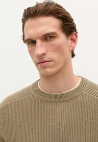 Knit sweater in muted olive green, featuring a ribbed texture and round neckline, worn over a cream-colored shirt.