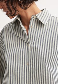 Button-up shirt with vertical blue and white stripes, featuring a spread collar and soft, textured fabric. Two buttons visible on the placket.