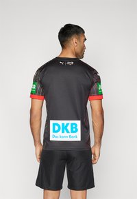 Črni atletski dresek z rdečimi poudarki in diagonlnimi rumenimi črtami, z logotipom "DKB" na hrbtni strani. V kombinaciji s črnimi kratkimi hlačami.