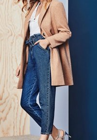 Cappotto in misto lana beige sopra una maglietta bianca e jeans in denim blu a vita alta. Sandali con tacco argentati completano l'outfit. Texture lisce, design sartoriale.
