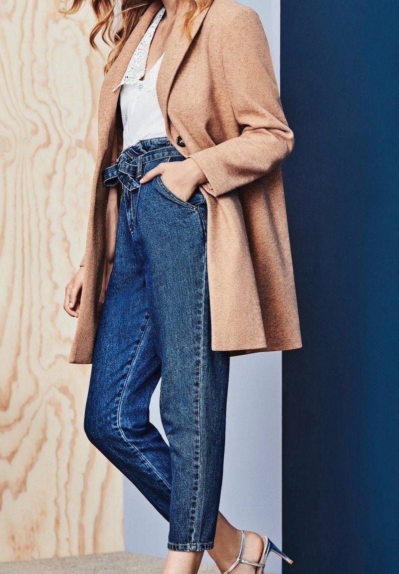 Cappotto in misto lana beige sopra una maglietta bianca e jeans in denim blu a vita alta. Sandali con tacco argentati completano l'outfit. Texture lisce, design sartoriale.