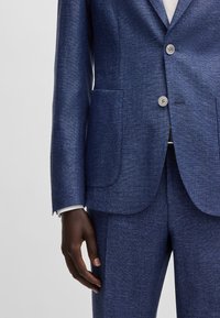 Blazer in lino blu navy con una finitura testurizzata, due tasche a toppa e bottoni argentati. I polsini mostrano una camicia bianca che sbuca.