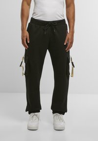 Mann iført svarte cargo sweatpants med beige stroppedetaljer og hvite sneakers, står mot en lys grå bakgrunn.
