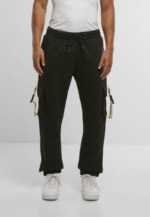 Mand iført sorte cargo-sweatpants med beige stropdetaljer og hvide sneakers, stående mod en ensfarvet lysegrå baggrund.