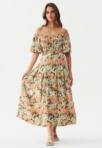 Bloemrijke off-the-shoulder maxi-jurk in bruin, met levendige oranje, groene en witte patronen, en een uitsnijdetail bij de taille.