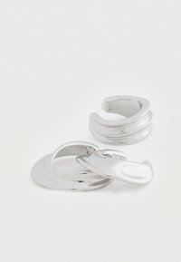 Dvoje lesklých strieborných kovových náušníc, jedny v krútenom manžetovom štýle a jedny v zloženom abstraktnom krúžku, na jednoduchom svetlom pozadí.