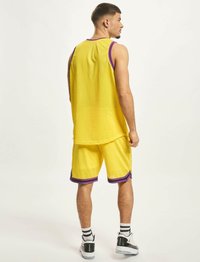 Geltonas akių krepšinio marškinėliai ir šortai su violetiniais akcentais. Sportinis kirpimas. Dėvimi su juodais ir baltais sportbačiais bei dryžuotomis kojinėmis.