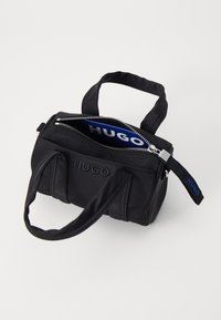 Sac à main en tissu noir avec deux poignées, fermeture éclair, logo "HUGO" en relief, et un intérieur bleu contrastant avec le texte "HUGO".