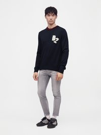 Jeune homme debout portant un sweat-shirt noir avec le logo "D2", un jean gris clair usé, des chaussettes blanches et des baskets noires à rayures blanches.