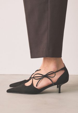Piedi con décolleté nere a punta con cinturini alla caviglia legati a fiocco, abbinati a pantaloni corti marrone scuro.