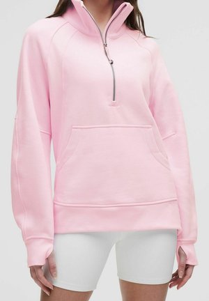 Kobieta ubrana w jasnoróżową bluzę z krótkim zamkiem i kieszenią typu kangur oraz mankietami na kciuki, w parze z białymi dopasowanymi szortami.