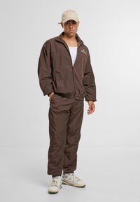 Moški s pegicami, oblečen v rjavo trenirko, belo majico brez rokavov, bež kapo in bele superge, stoji z rokami v žepih pred enotno ozadje.