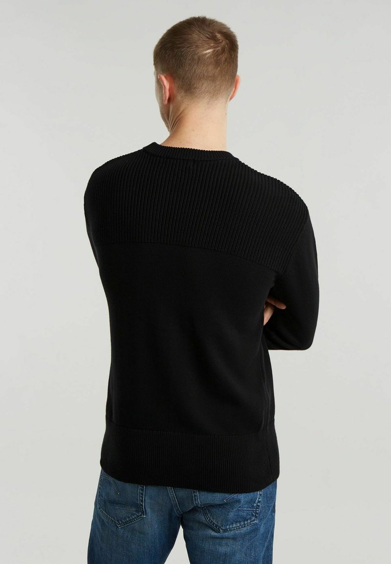 Suéter negro de punto con textura acanalada en la parte superior de la espalda y las mangas, combinado con jeans azules. Diseño sencillo y ajustado.