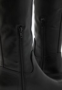 Black leather knee-high boots with side zippers, smooth texture, and reinforced stitching. Features a minimalist design and durable hardware.