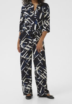 Femme portant une combinaison noire, bleue et blanche à motif abstrait avec manches retroussées et ballerines noires, debout devant un fond uni.