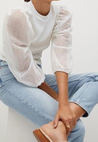 Weißer strukturierter Pullover mit transparenten Puffärmeln; kombiniert mit hellblauen Jeans. Das Model sitzt und zeigt einen braunen Sandalen-Schuh an einem Fuß.
