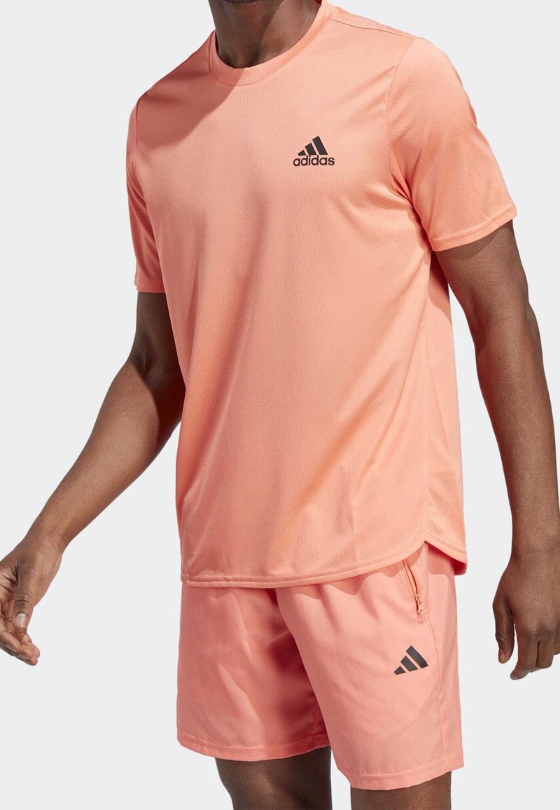 Camisa atlética de color coral claro y pantalones cortos de tela transpirable. La camisa tiene un logo negro de Adidas en el pecho; los pantalones cortos cuentan con un bolsillo con cremallera.