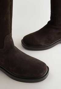 Botine din suede maro, cu o textură netedă, vârf ușor rotunjit și talpă plată din cauciuc. Prezintă cusături discrete de-a lungul cusăturilor.