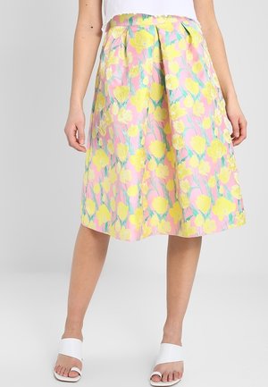 Femme portant une jupe trapèze rose à imprimé floral jaune, associée à des sandales blanches ouvertes et un haut blanc, debout devant un fond uni.