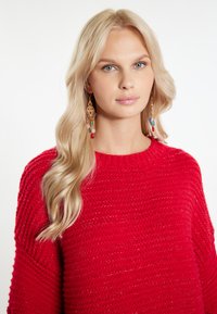 Roter Strickpullover mit strukturiertem Finish und weitem Ausschnitt. Das Modell trägt farbenfrohe, hängende Ohrringe mit Perlen und metallischen Akzenten.