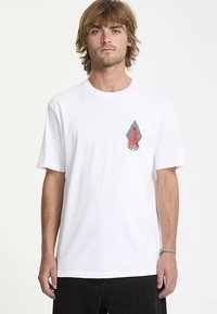 Maglietta bianca in cotone con grafica di una figura rossa su uno sfondo a rombi blu e nero, con maniche corte e vestibilità comoda.