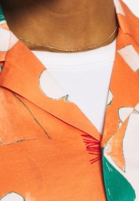 Koszula z krótkim rękawem z pomarańczowej tkaniny z abstrakcyjnymi białymi i zielonymi kształtami, z teksturowanym kołnierzem i warstwową białą podkoszulką.