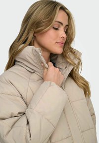 Beige vadderad jacka med hög krage, kviltad design och dragkedja. Mjuk textur med subtil glans.