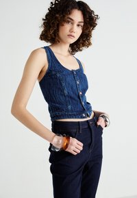 Top corto in denim di colore blu scuro con un motivo a texture, dotato di chiusura a bottone e spalline larghe, abbinato a jeans blu scuro e bracciali sovrapposti.