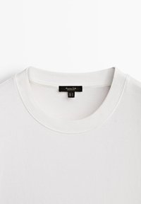 T-shirt en coton blanc présentant un col rond côtelé, une texture lisse et une étiquette noire à l'intérieur du col indiquant la taille et les instructions d'entretien.