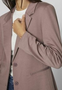 Veste légère mauve avec une coupe sur mesure, fermeture à un bouton et deux poches avant. Texture lisse avec un col structuré et des manches.