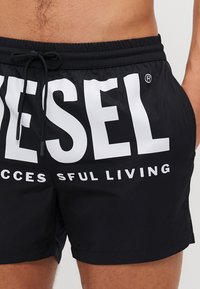 Sorte badetights med snor i livet, med store hvite "DIESEL"-trykk og teksten "SUCCESSFUL LIVING". Glatt, lett materiale.