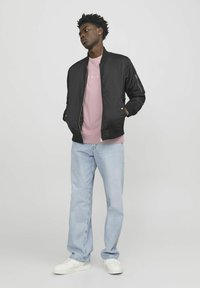 Schwarze Bomberjacke mit einem Reißverschluss vorne, getragen über einem rosa T-Shirt. Hellblaue Jeans und weiße Sneaker runden das Outfit ab.