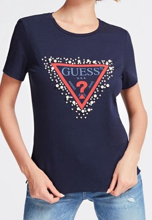 Laivastonsininen t-paita, jossa koristeltu GUESS-logon kolmio punaisena ja sinisenä, pukeutuneena sinisiin farkkuihin, kädet säätävät helmaa.