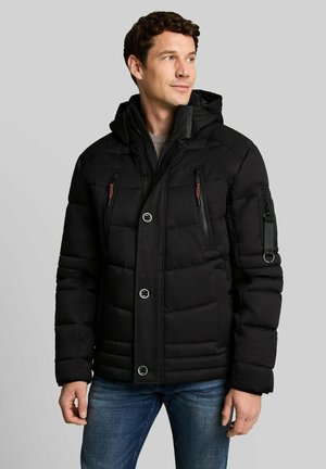 Man wearing a black quilted winter jacket with multiple zippered pockets and blue jeans, looking to the side.